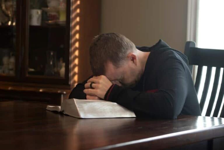 a man sitting at a table hands folded on a bible featured image for 8 Tips for Trusting God in Difficult Times To Stand Strong in faith
