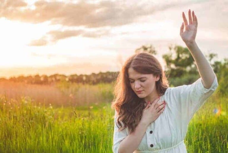 a woman on her knees in a field hand raised high praying How the Power of Prayer Changes Things