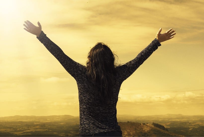 an image of a woman with both arms stretched towards the sky overlooking a golden sunset over a mountain featured image for What Does the Bible Say About Peace?