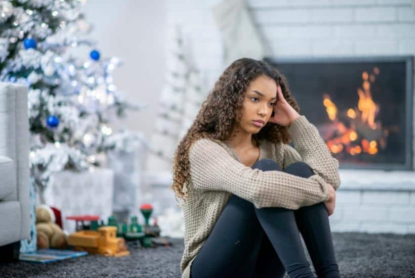 a sad woman wearing blue jeans and beige sweater sitting on the floor with a Christmas tree in the background featured image for Best Books About Grief For Surviving The Holidays