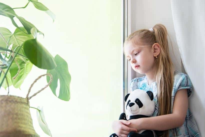 a little girl sitting on a ledge looking out the window holding a panda bear looking sad featured image for 13 Heart Warming Grief Books For Children
