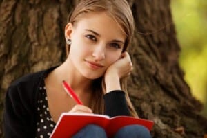 a young woman sitting against a tree with a red pen in her hand and a red notebook featured 22 Guided Grief Journaling Prompts