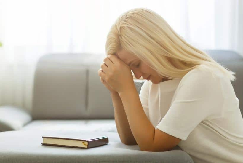 a blonde haired woman on her knees, head bowed, hands folded in prayer with a bible laying on a beige couch in front of her featured image for Trusting God in the Midst of Grief: 7 Helpful Tips
