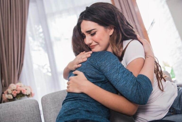 a woman wearing a blue shirt comforting another friend sitting on a beige sofa featured image for What Not to Say to Someone Grieving: 16 Accurate Tips