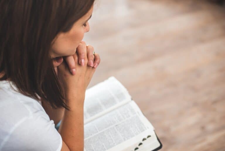 a woman in a white shirt folded hands in prayer with her elbows on her opened bible.featured image for Perspective In Christianity 20 Best Verses For New Believers